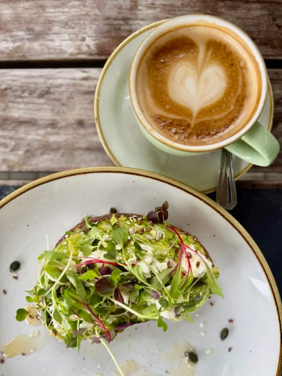 Latte with heart-shaped latte art and avocado toast topped with microgreens and seeds on rustic wooden table