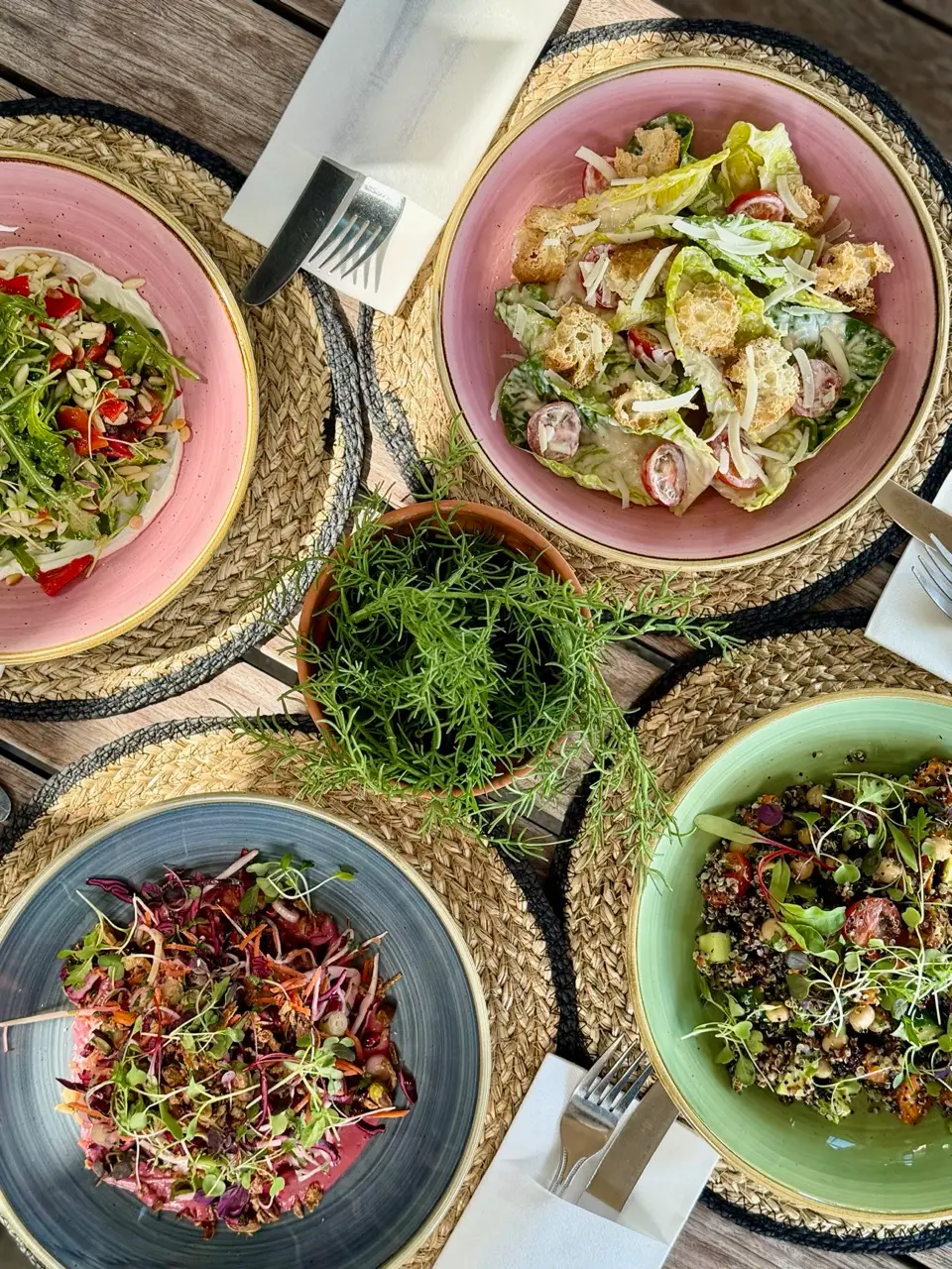 Four colorful bowls filled with fresh salads and greens on woven placemats, displayed on a wooden table with cutlery and a small herb plant in center.
