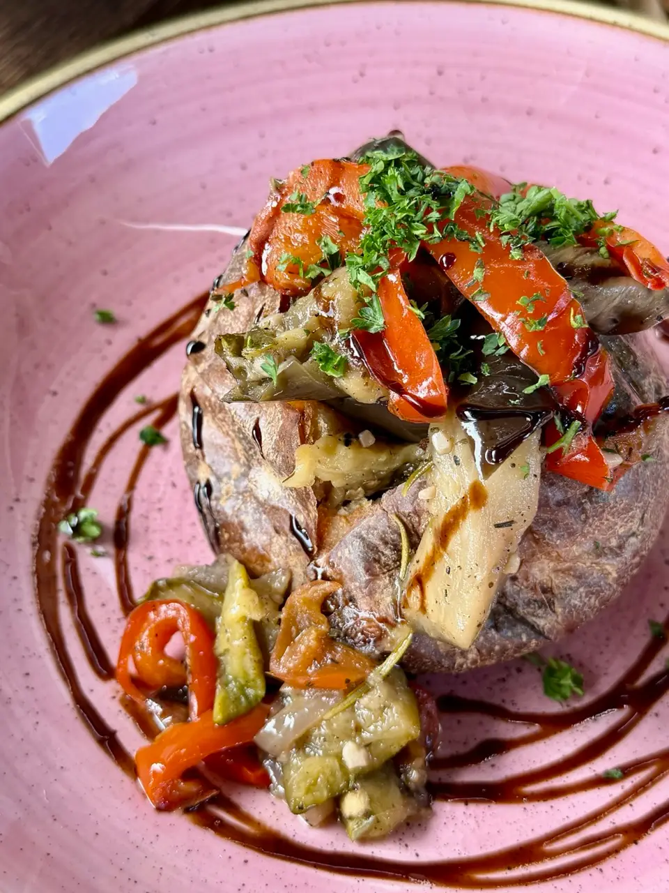 Baked potato topped with sautéed peppers, eggplant, herbs, and drizzled balsamic glaze on pink plate