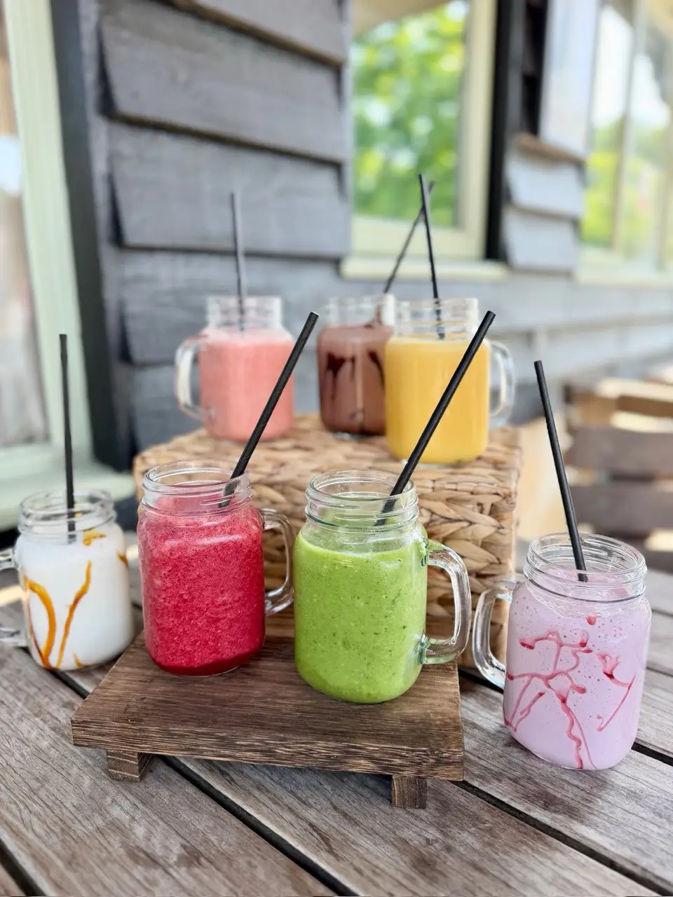 Seven colorful smoothies in glass jars with black straws on a wooden table outdoors