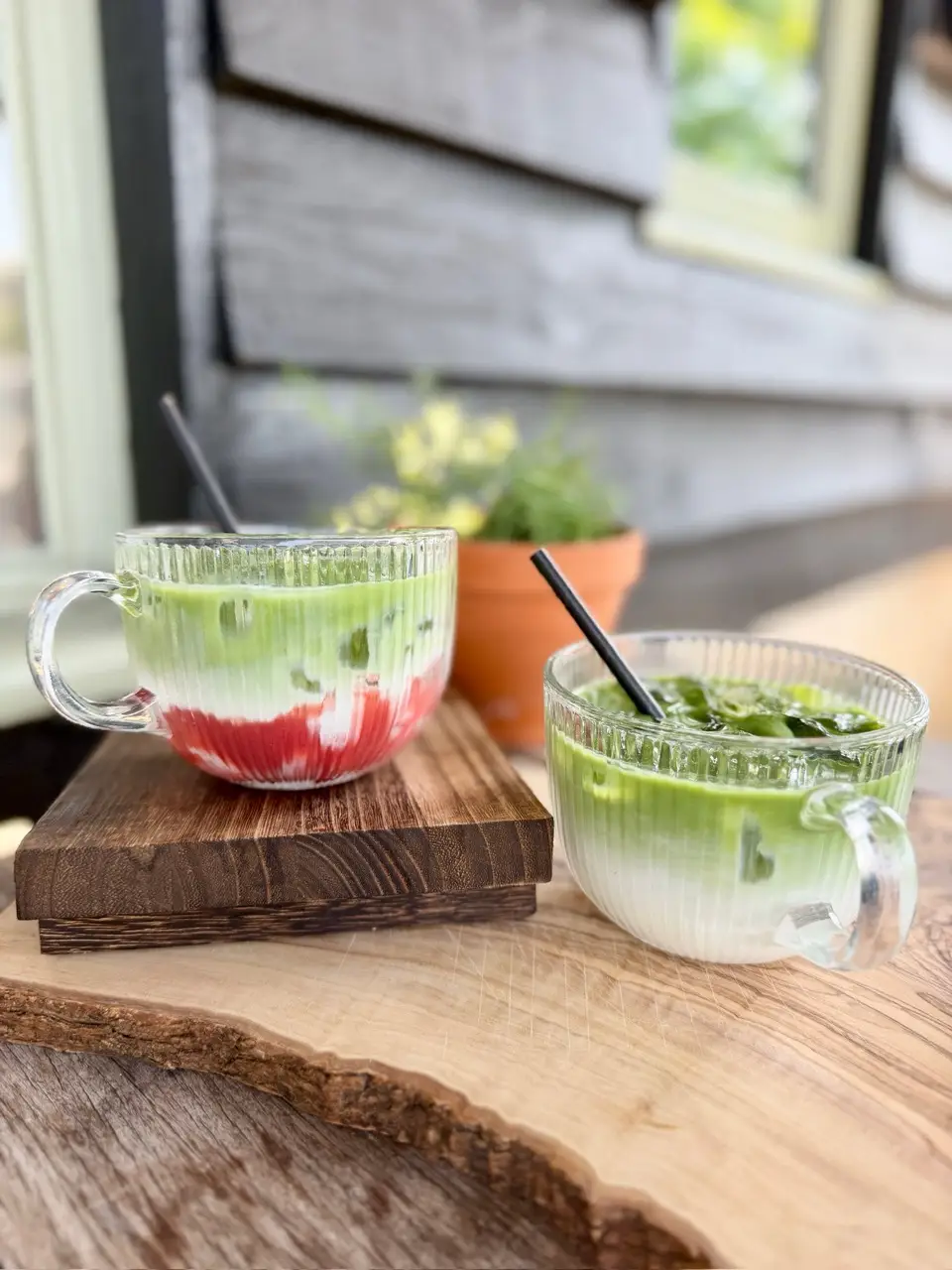 Two glass cups with iced green matcha drinks on wooden surfaces, one with red syrup layering and black straws, rustic background with potted plant.