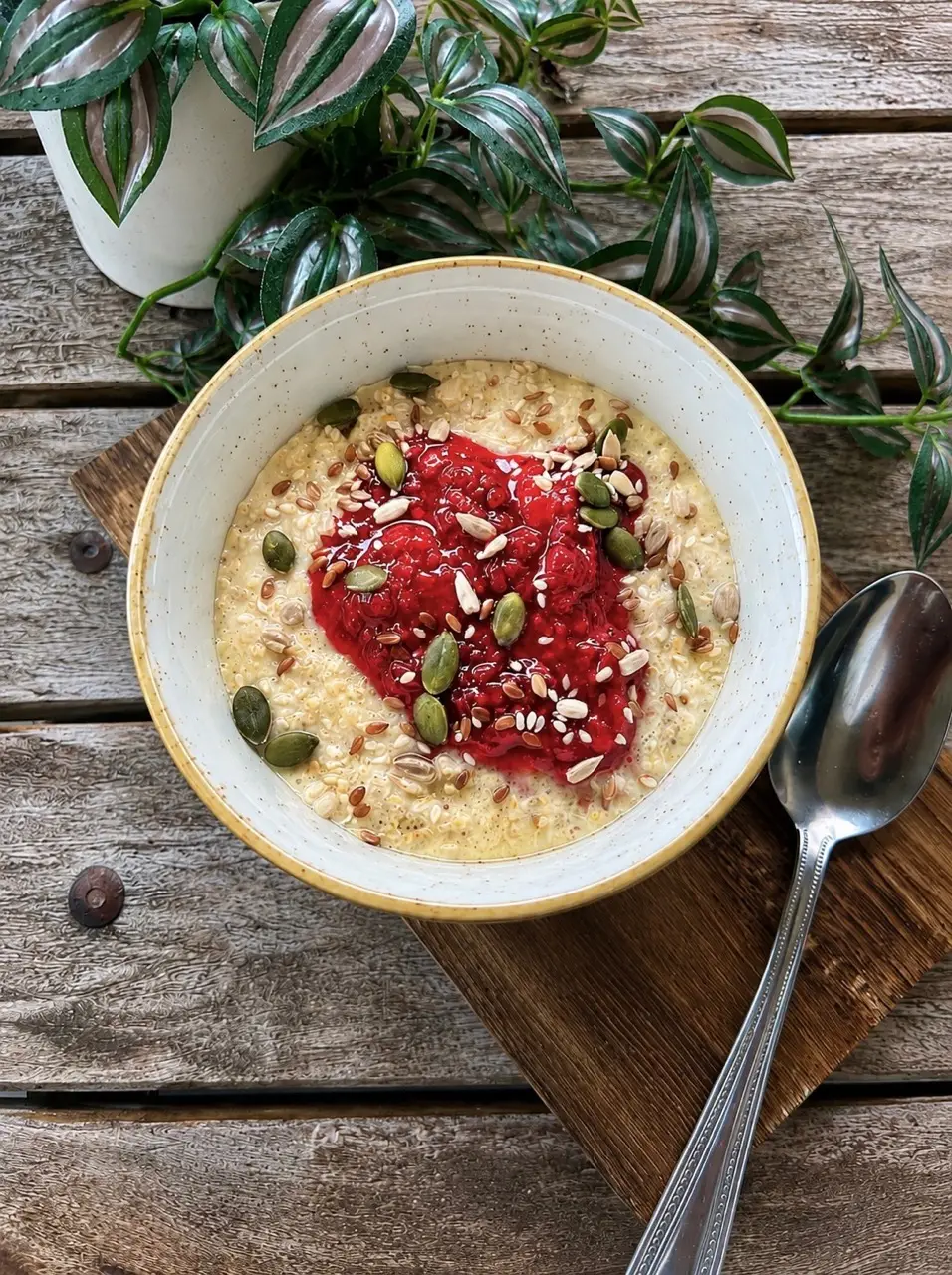 Bowl of porridge topped with red berry compote and seeds on wooden table with spoon and potted plant