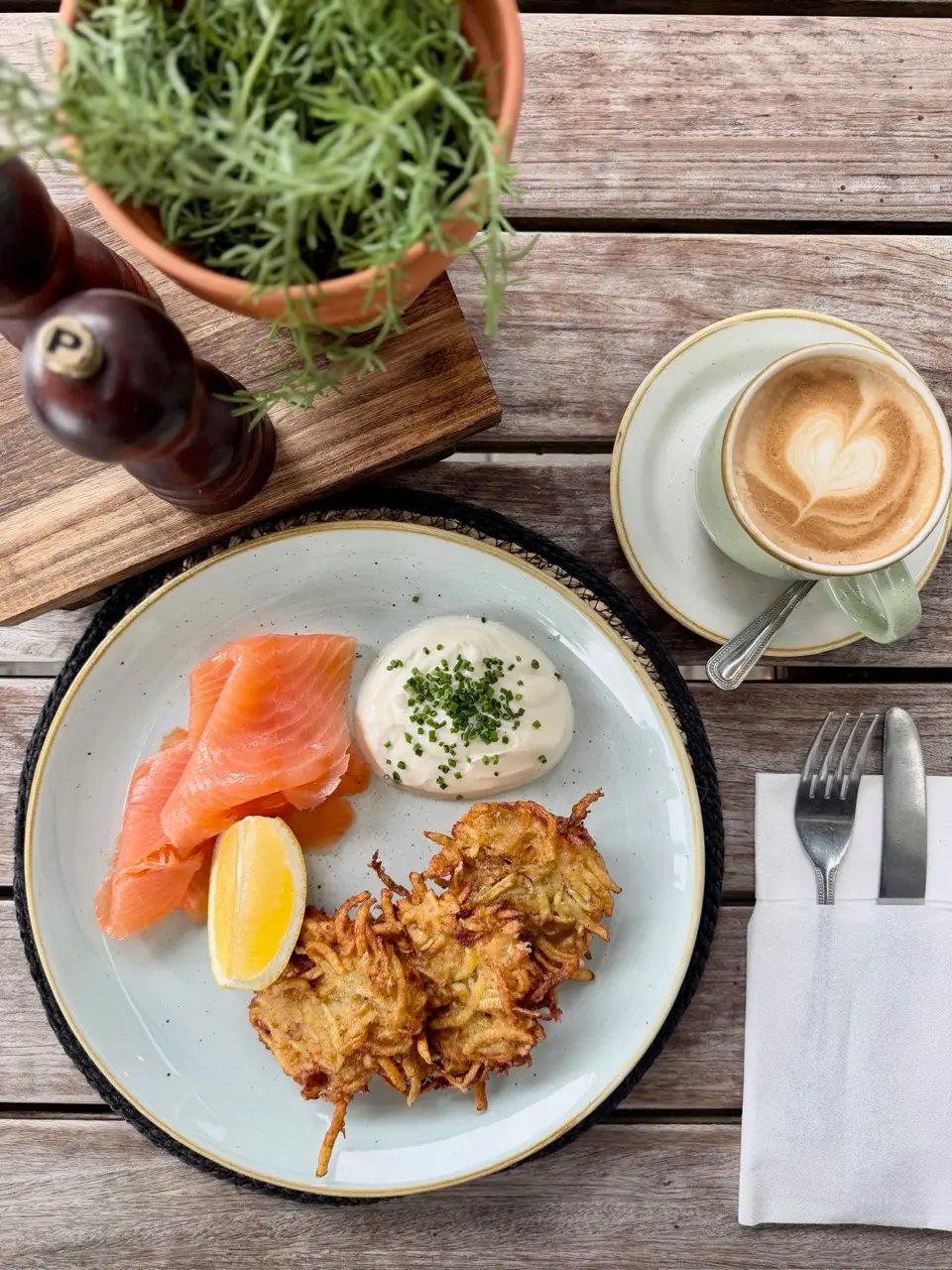 Plated smoked salmon, potato latkes with sour cream and chives, lemon wedge, coffee with heart foam art on wooden table