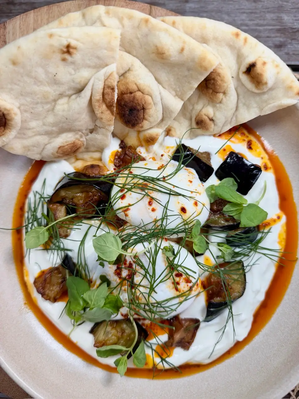 Two poached eggs on creamy sauce with grilled aubergine, herbs, spices, and flatbreads on a beige plate