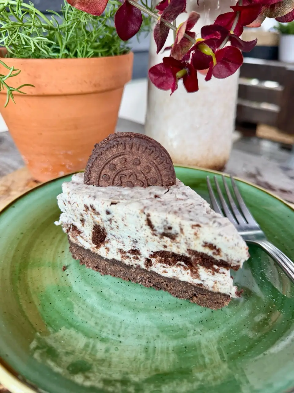 slice of cookies and cream cheesecake on a green plate with a fork, plant pot and flowers in background