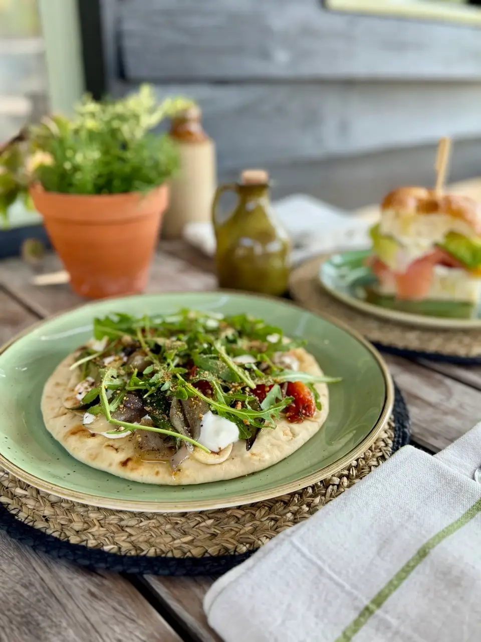 Flatbread topped with greens, mushrooms, and sauce, with a sandwich and condiments on a wooden table with a potted plant in background