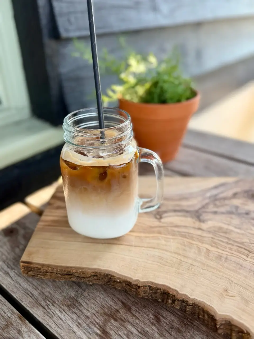iced latte in a glass jar with a black straw on a wooden board with a plant pot in background