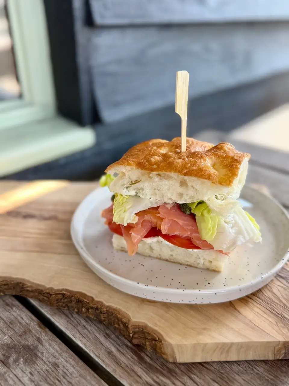 Sandwich with smoked salmon, lettuce, and tomato on focaccia bread on a plate and wooden board