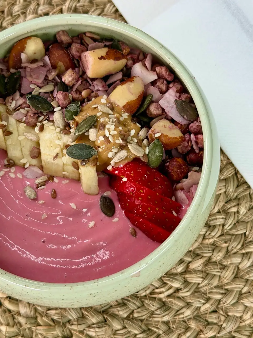 Pink smoothie bowl topped with banana slices, strawberries, nuts, seeds, and peanut butter in a bowl on woven mat